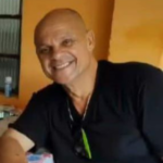 Retrato de Antonio Rubens Veloso sorrindo para a câmera. Ele está usando uma camiseta preta e aparece em um ambiente interno com iluminação quente, transmitindo uma expressão acolhedora e descontraída.