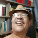 Retrato de Benedito de Almeida Sampaio Filho sorrindo para a câmera. Ele usa chapéu marrom e óculos, e está em um ambiente interno com estante de livros ao fundo, transmitindo uma expressão alegre e acolhedora.