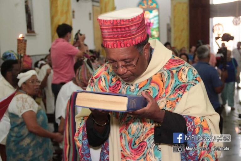 Participante da Celebração Inculturada Afro-brasileira de Itu veste trajes tradicionais coloridos e segura um livro junto ao rosto em um gesto de reverência. Ao fundo, outras pessoas acompanham a celebração em um espaço comunitário, destacando a espiritualidade e a valorização das tradições afro-brasileiras.