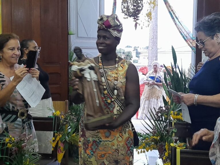 Pessoa usando vestimenta e adereços inspirados nas tradições afro-brasileiras participa de uma cerimônia cultural em ambiente interno. Ao redor, outras pessoas acompanham a atividade e registram o momento, enquanto elementos decorativos reforçam o caráter de celebração e valorização da cultura negra.