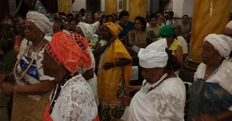Grupo de mulheres negras participa de uma celebração afro-brasileira, vestindo roupas e turbantes tradicionais em diferentes cores. Elas caminham em procissão enquanto o público acompanha a cerimônia, em um momento de espiritualidade, memória e valorização das tradições afro-brasileiras.