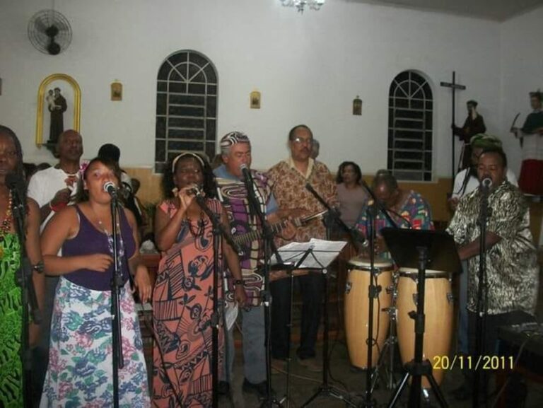 Integrantes do Coral da UNEI realizam uma apresentação musical em um espaço interno. O grupo canta acompanhado por instrumentos de percussão e sopro, reunindo homens e mulheres em uma celebração da cultura e da música afro-brasileira.