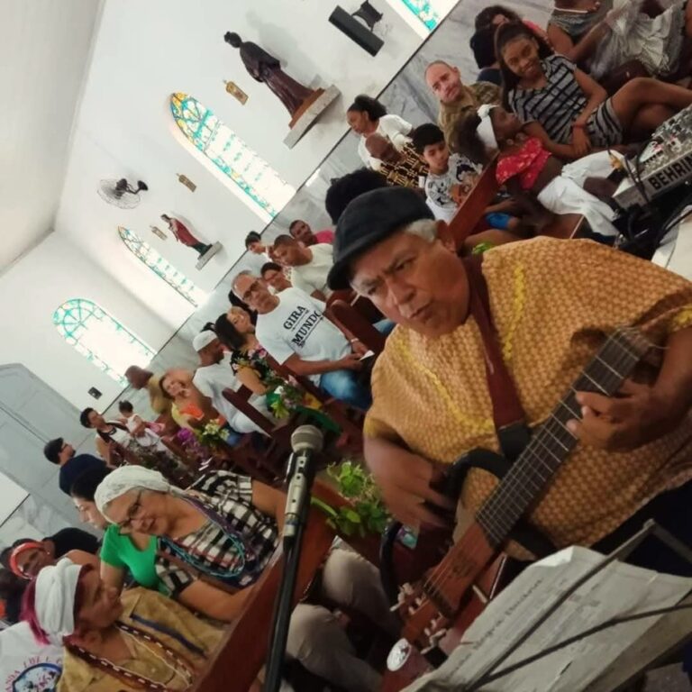 Integrantes do Coral da UNEI realizam uma apresentação musical em um espaço de celebração. Em primeiro plano, um músico toca violão ao lado de outros instrumentistas e cantores, enquanto o público acompanha a atividade em um ambiente de convivência, espiritualidade e valorização da cultura afro-brasileira.