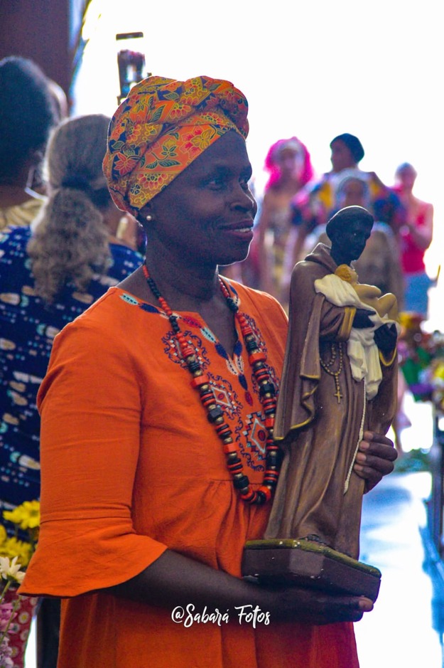 Mulher negra veste roupas e turbante em tons de laranja e segura uma imagem religiosa durante uma celebração afro-brasileira. Ela usa colares de contas e sorri discretamente, enquanto outras pessoas acompanham a cerimônia ao fundo em um ambiente de fé e valorização das tradições culturais.