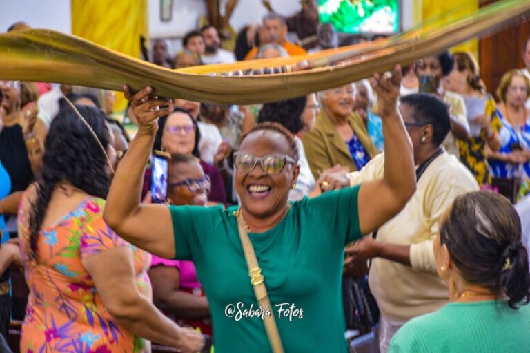 Mulher negra sorridente levanta um tecido colorido durante uma celebração cultural. Ao redor, diversas pessoas participam da atividade em um ambiente festivo, marcado pela música, pela dança e pela valorização das tradições afro-brasileiras.