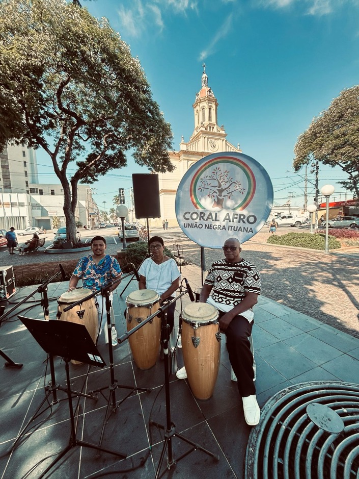 Integrantes do Coral Afro Nego Fugido se apresentam ao ar livre em uma praça. Três percussionistas tocam tambores em frente a um banner do grupo, tendo ao fundo uma igreja histórica e a paisagem urbana, em um momento de celebração da cultura afro-brasileira.