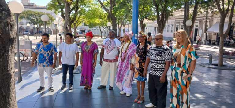 Integrantes do Coral da UNEI reunidos ao ar livre em uma praça. O grupo, formado por homens e mulheres de diferentes idades, está alinhado lado a lado, vestindo roupas com estampas afro-brasileiras e trajes tradicionais, em um momento de convivência e valorização da cultura negra.