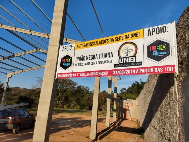 Faixa da UNEI instalada em uma estrutura ao ar livre, divulgando o projeto 'Cursinho Popular da UNEI'. O banner apresenta informações sobre inscrições, parceiros e apoiadores, tendo ao fundo árvores e o céu azul.