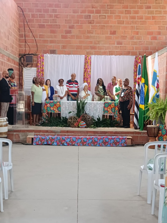 Participantes reunidos na sede da UNEI durante uma cerimônia cultural. Um grupo de pessoas está posicionado à frente de um palco decorado com tecidos coloridos, bandeiras e elementos da cultura afro-brasileira, enquanto o público acompanha a atividade em um ambiente de celebração, memória e valorização da cultura negra.