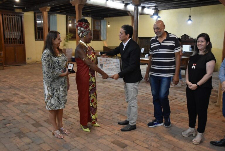 Representantes da UNEI participam de uma cerimônia de homenagem em um espaço cultural. No centro da imagem, uma pessoa com vestimenta de inspiração afro-brasileira recebe um cumprimento e uma lembrança simbólica, enquanto outras pessoas acompanham o momento de reconhecimento e celebração.
